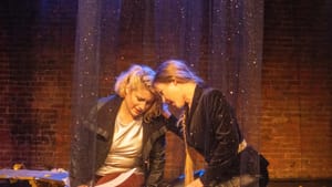 Two white women actors lean heads tenderly together, inside a glittery curtain. They wear a mix of leather, velour, & satin