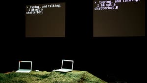 Two laptops in front of a black wall. Hanging screens read: "typing and talking. I am not a chatterbot."