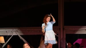 Jenna Kuerzi, a white woman, as Alice in a blue dress & white apron. She poses on a table with ensemble members around her