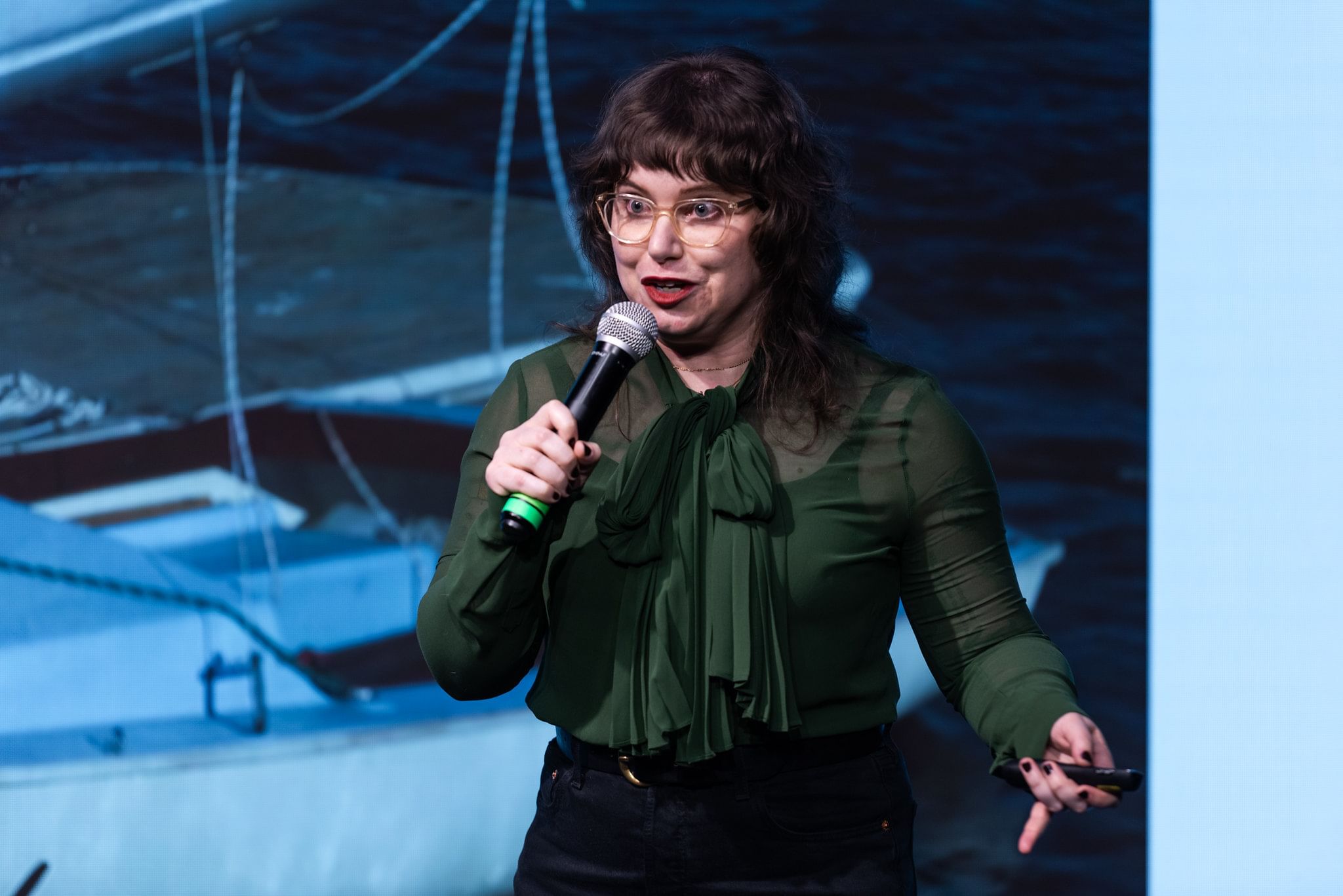 Zeidman holds a microphone on stage, dressed in mostly black, an image projected on the screen behind her