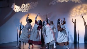 Four women, wearing drab dresses with puffed sleeves and white aprons, extend one arm upward in a dance.