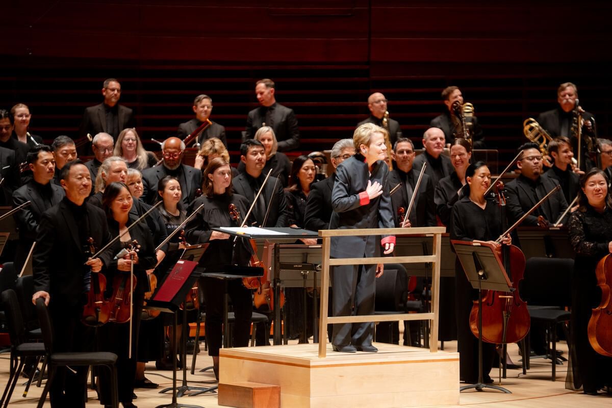 Alsop, at the conductor’s podium with orchestra standing around her, faces the audience, smiling with hand on her heart.
