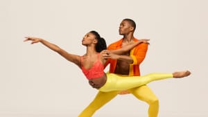 Two Black dancers in reds tops, yellow leotards, looking to their left, one standing on their feet while carrying the other