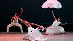 Four dancers in various poses on stage, wearing all white. One holds an umbrella open.