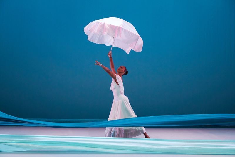 Stamatiou, a tall Black woman, poses with limbs outstretched in an airy white dress, holding a white umbrella aloft.