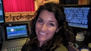 Selfie of Docherty in a booth with lighting consoles and monitors behind her, a stage can be seen through a window