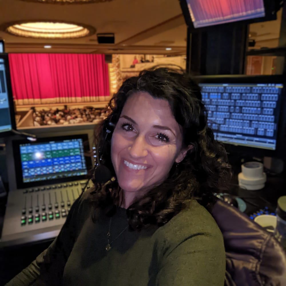 Selfie of Docherty in a booth with lighting consoles and monitors behind her, a stage can be seen through a window