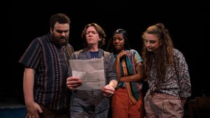 Four people on stage in casual wear stand together, looking at a white sheet of paper that looks like a bill