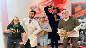 Five people in various outfits, including one in a lab coat; a Nirvana shirt; dresses—pose in front of a 'Red Rum; neon sign