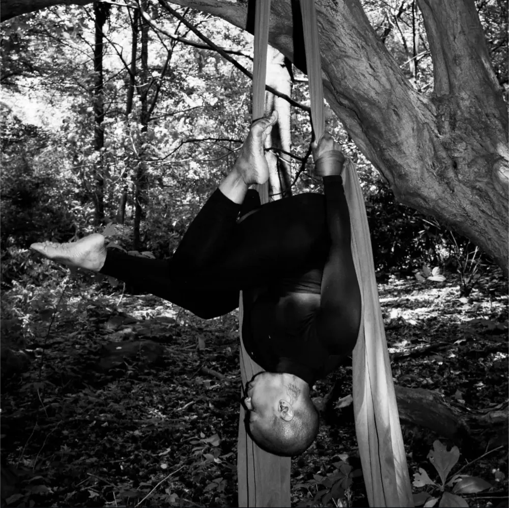 Black and white photo of a performer outdoors hanging from a tree via an aerial silk