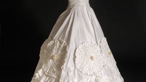 Mannequin wears the silky white short-sleeve gown, with giant complex rosettes & luxurious pleated layers on the skirt.