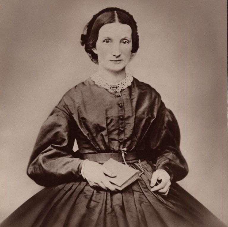 Historic sepia photo of Preston, a sober white woman in a dark dress with puffed sleeves & a full skirt. She holds a book.