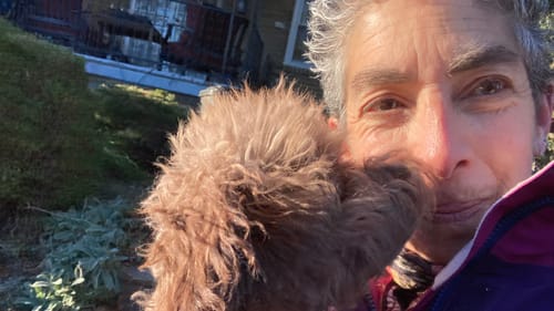 Close-up selfie of a tiny, fluffy brown poodle nuzzling Anndee’s smiling face on a sunny day.