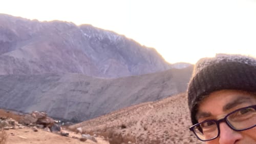 A selfie of Anndee, who smiles at the far bottom right corner of a photo of the high, sharp-edged Andes, against a golden sky