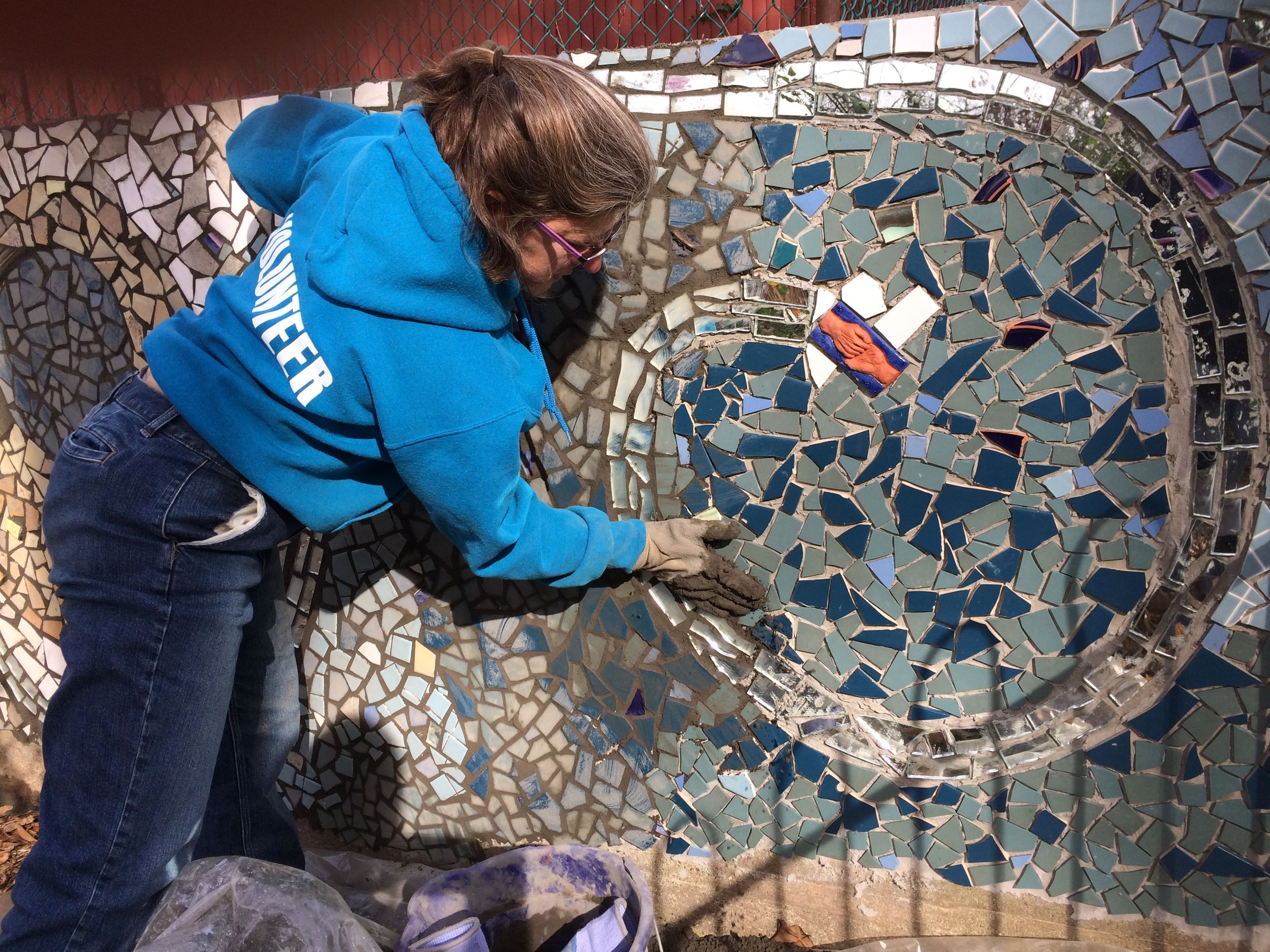 Elyssa, a white woman in a blue hoodie, leans over while working gray grout into a blue mosaic mural.