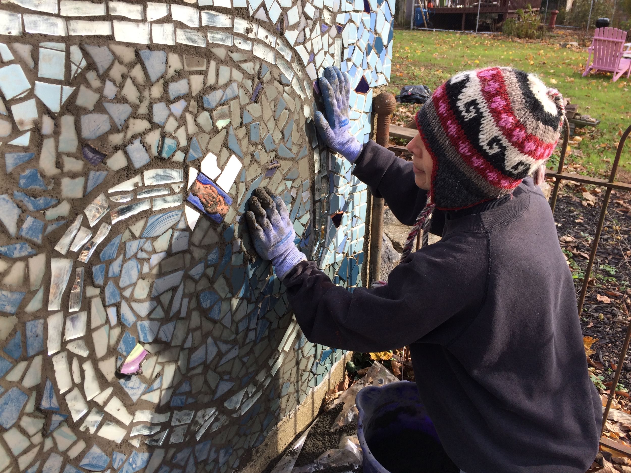 Anndee, a white woman in a patterned knitted hat wears gloves as she applies gray grout to a blue mosaic mural.
