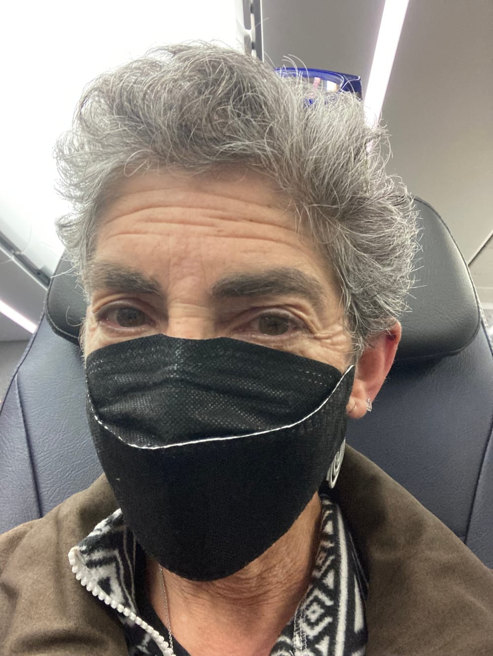 A selfie of Anndee, a white woman with short gray hair, wearing a black facemask in an airline cabin.