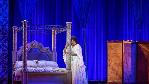 Harcum, a Black woman in an opulent lacy white nightgown, sings while clutching the gilded pillar of her 4-poster bed