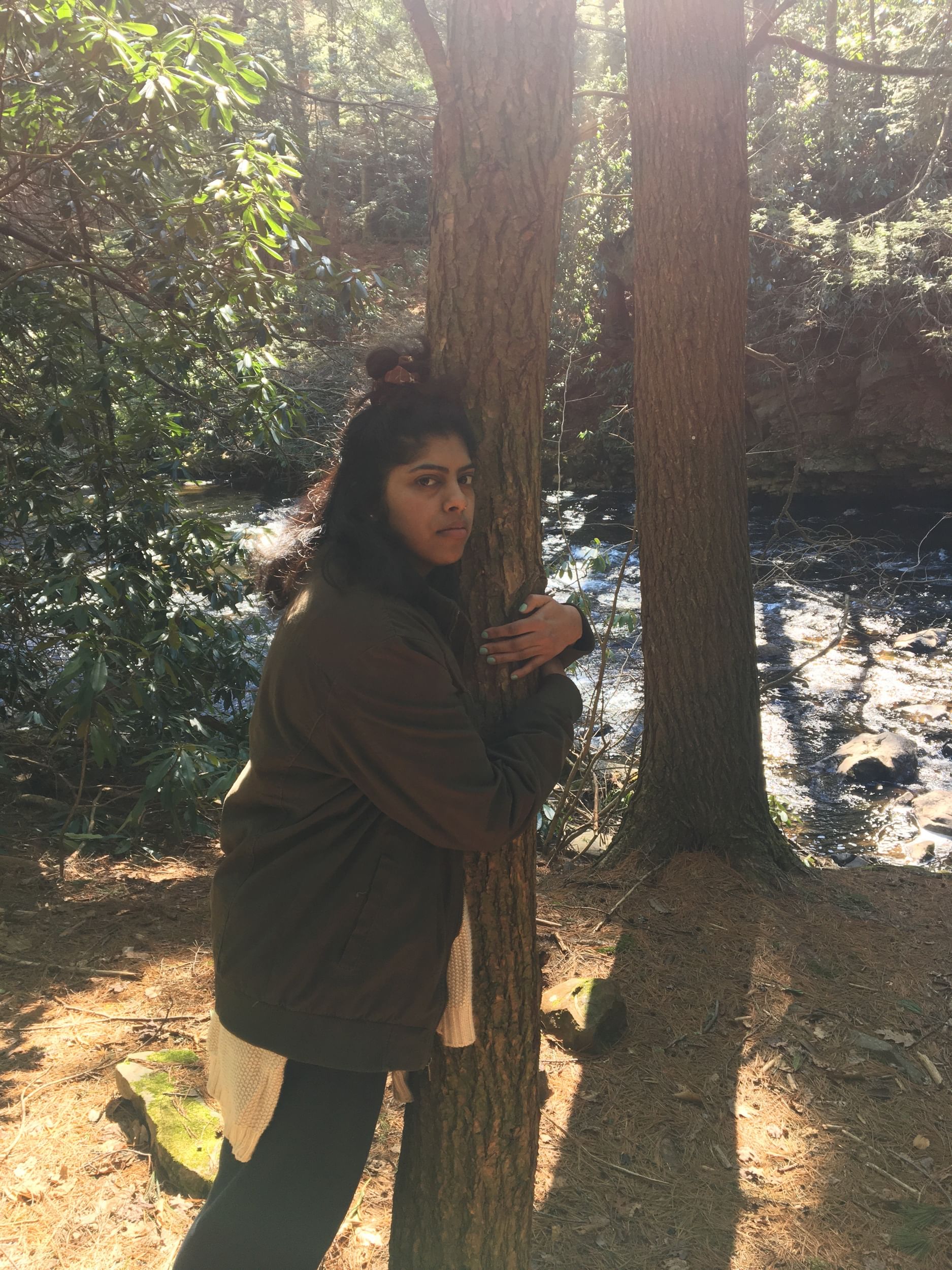 Christina stands embracing a slender tree beside the woodsy Wissahickon river. She has a serious expression.