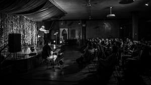 B&W wide shot, performance hall & a white bald man brandishing two foils in a crossed “X” position in front of an audience