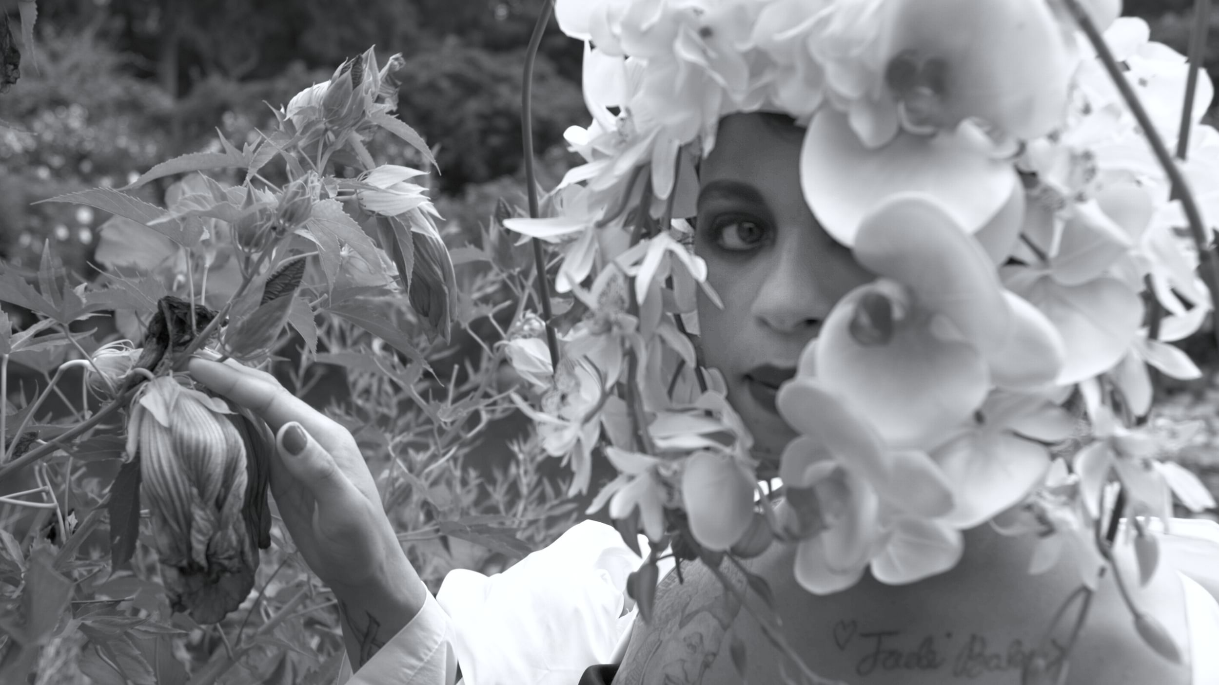 Lush black & white film still shows a Black woman encased in orchid-like flowers, stroking another flower with one hand.