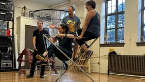 Three people balance delightedly on a small structure of metal chains and poles while Kennedy walks around them smiling.