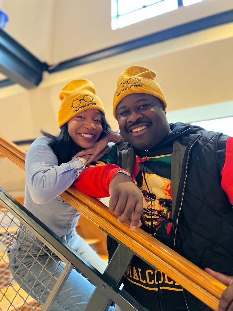 Nixon and Davis pose together smiling on the Arden’s lobby staircase, wearing yellow Ijames beanies.