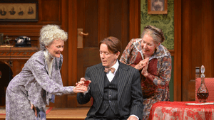 Two women in floral patterned gowns offer a drink in a glass to a man in a pinstriped suit.