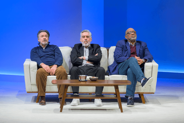 The three men sit facing outward on a pale midcentury modern couch. They seem resentful yet resigned.
