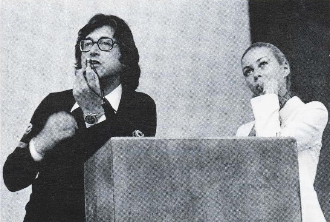 Black and white photo, Kane and a woman to his right at a podium. Kane speaks into a small mic, the woman looks to audience