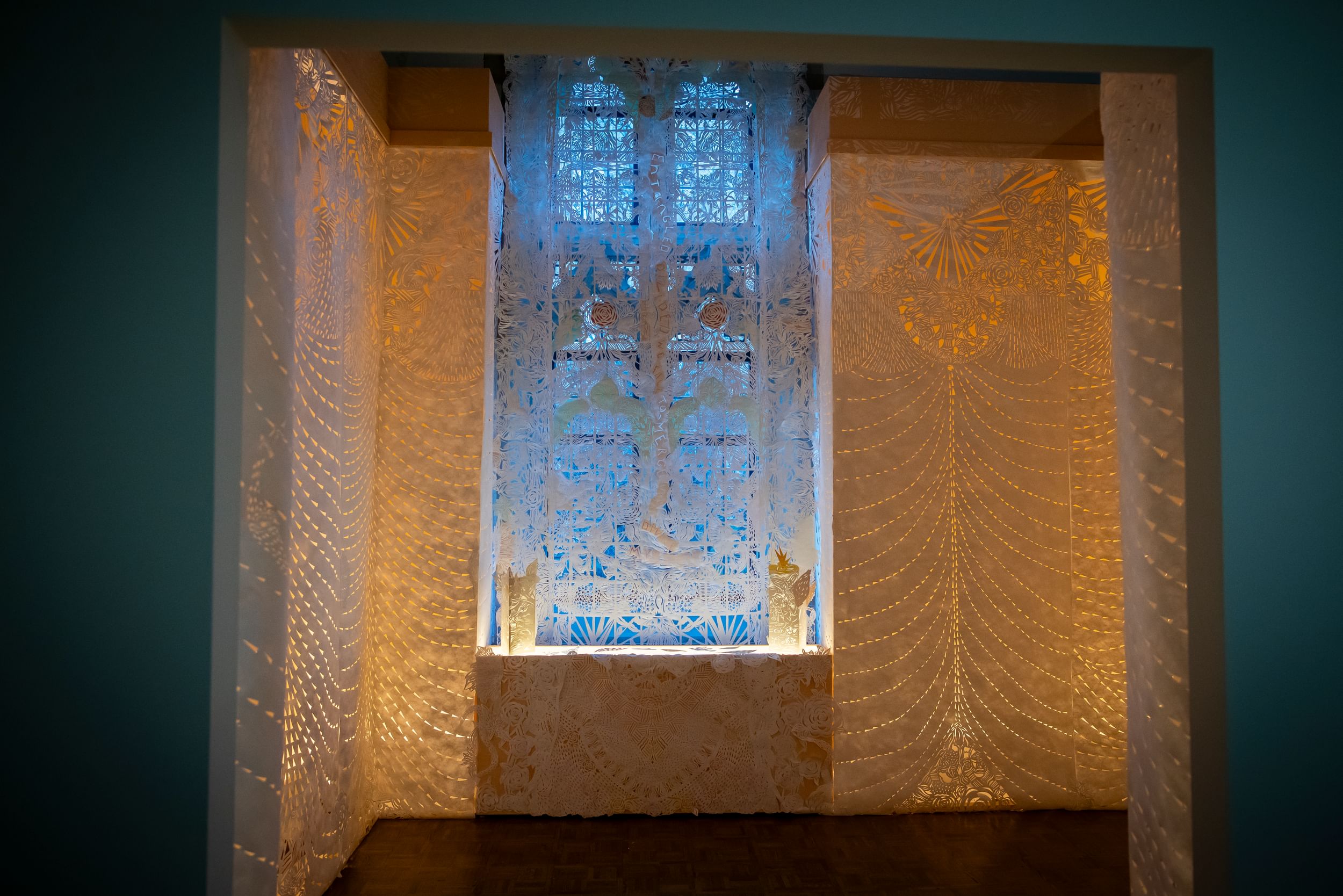 Pale wallpaper-like pieces in a windowed corner of the gallery seem to glow with lace-like filigrees of orange light