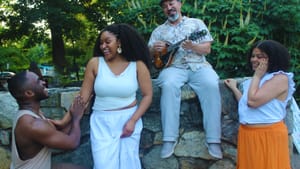 All 4 performers outside in a park smiling and laughing, looking towards Kanu’s character. Lindner holds a mandolin
