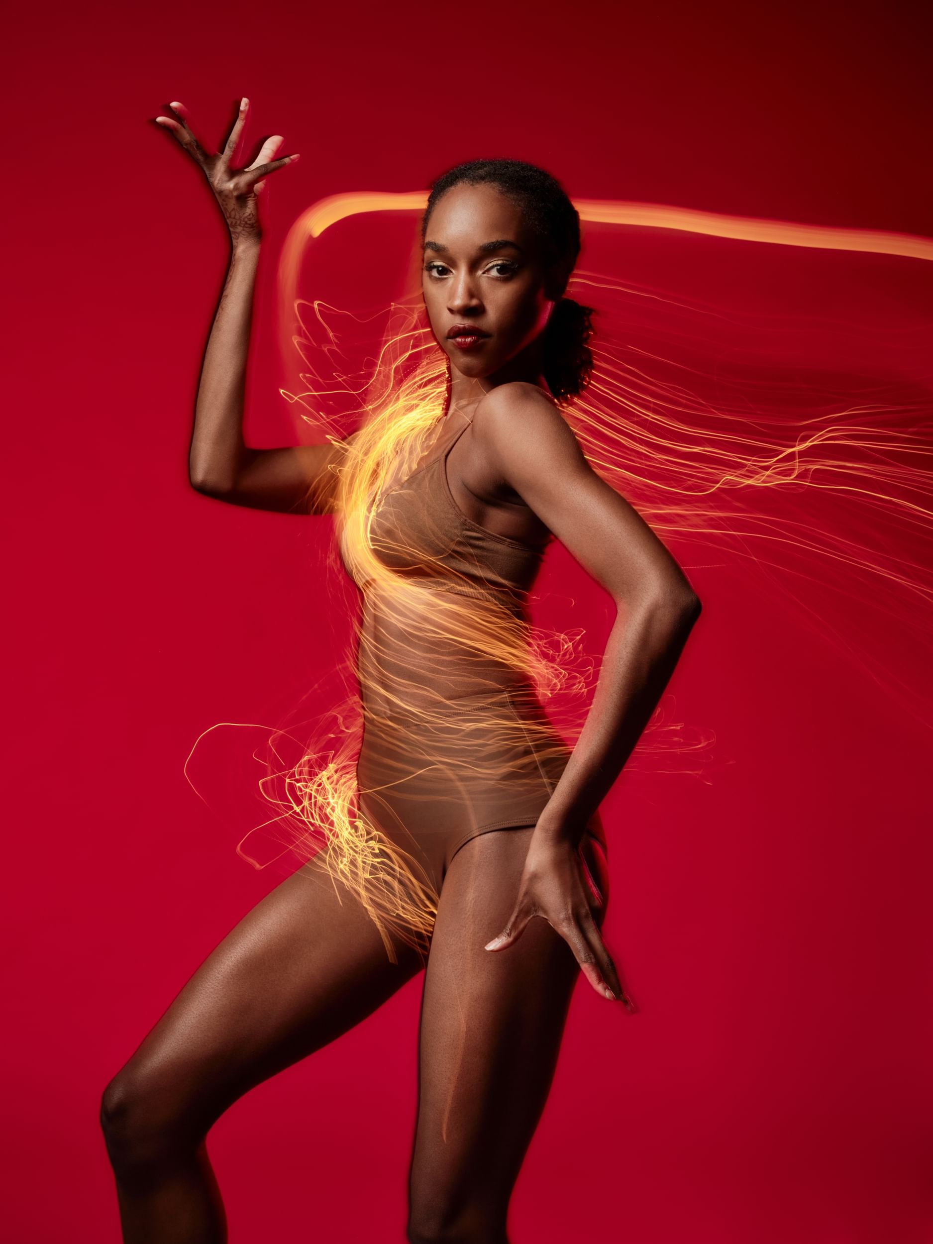 Simpson, a Black woman in skin-toned shorts & top, poses dynamically, right arm reaching up, wrapped in a yellow light effect