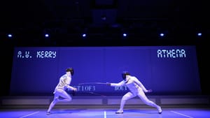 A scene from the play. Two female fencers in white protective uniforms lunge at each other on a blue-lit stage.