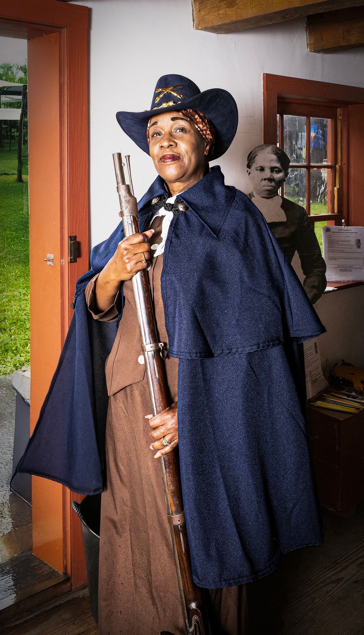 A Black woman in a 19th-century blue hat and cape stands holding a long gun in the doorway of a historic house.