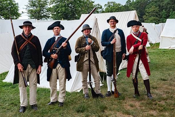 Five soldiers in historic 18th-century dress pose solemnly together with guns, in front of white tents.
