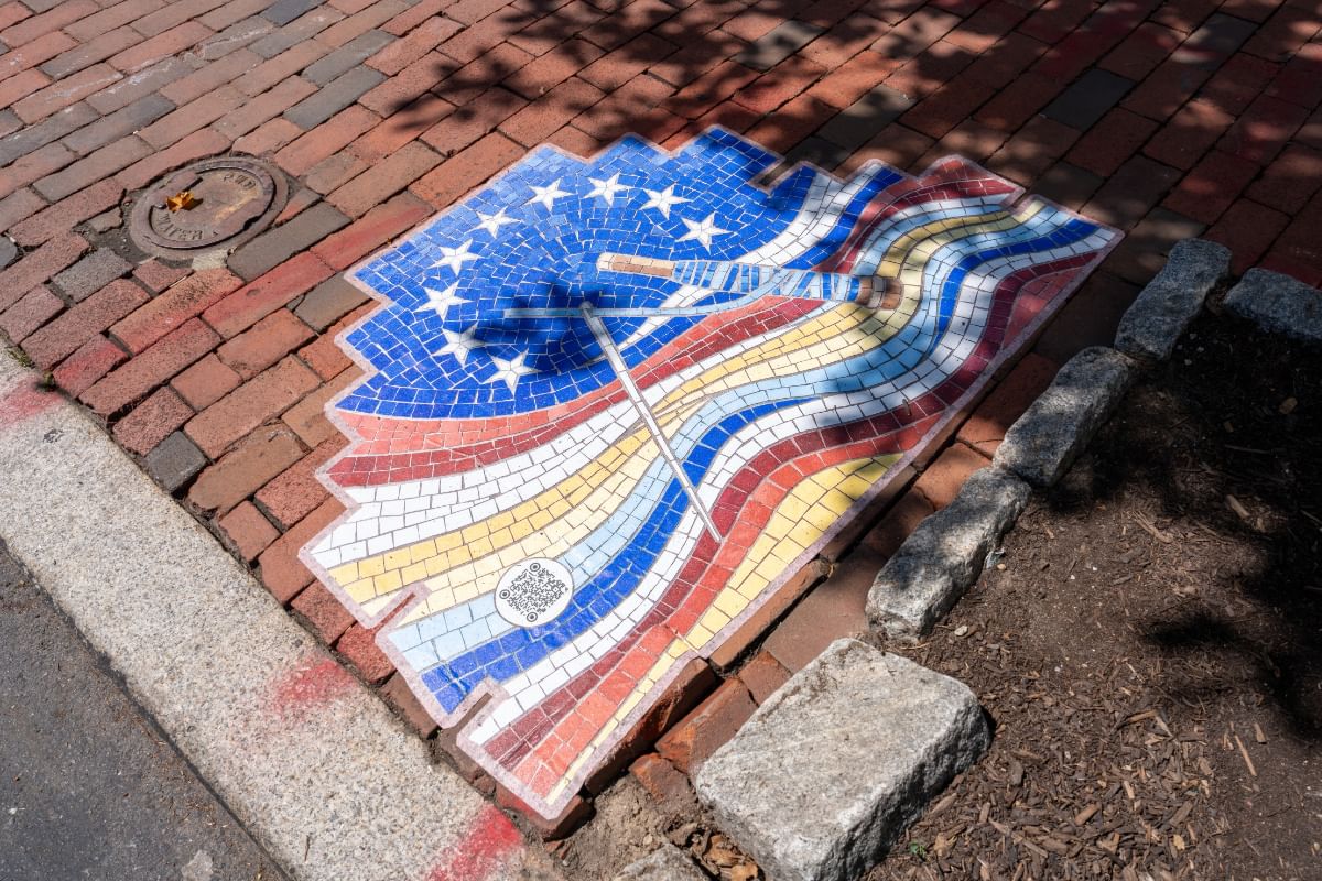Closeup on colorful mosaic-style art evoking the US flag on a brick pathway, centering a needle and skein of thread.