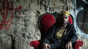 Cousar, a Black man dressed in black, sits sternly in a red upholstered chair in front of an old, graffitied stone wall.