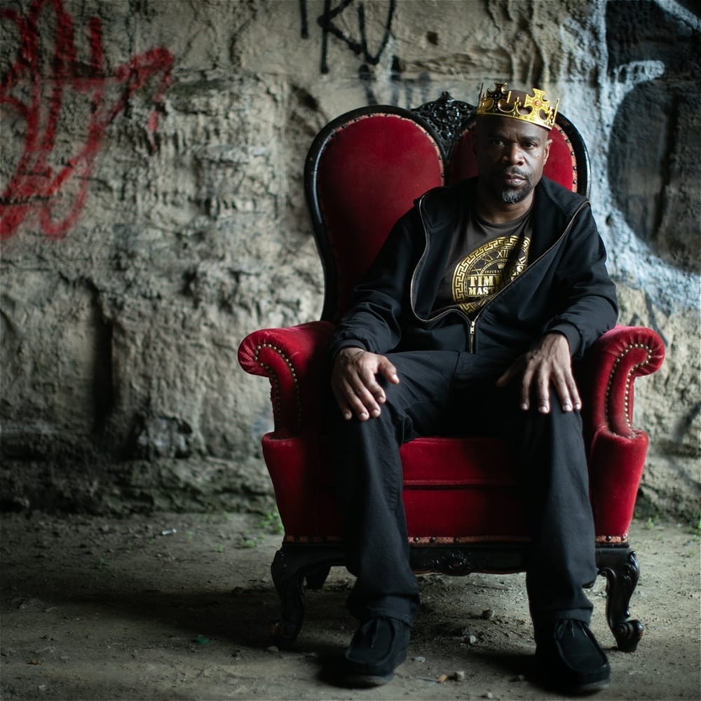 Cousar, a Black man dressed in black, sits sternly in a red upholstered chair in front of an old, graffitied stone wall.
