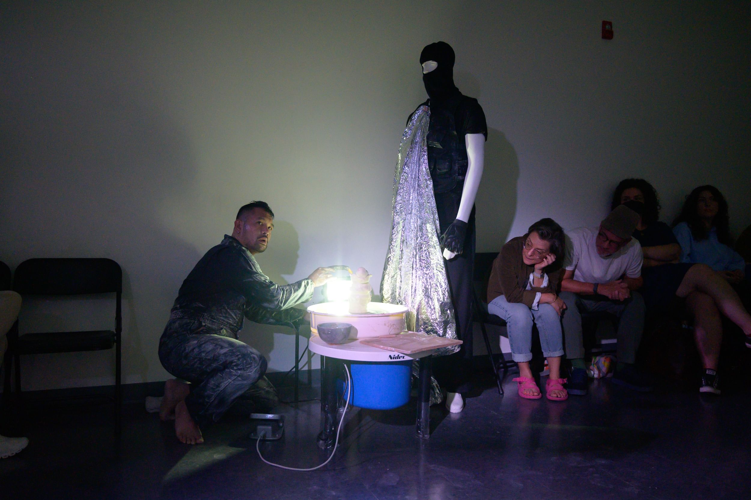 Jain, in a clay-smeared jumpsuit, squats and manipulates clay & light. A mannequin dressed like an ICE agent stands over him