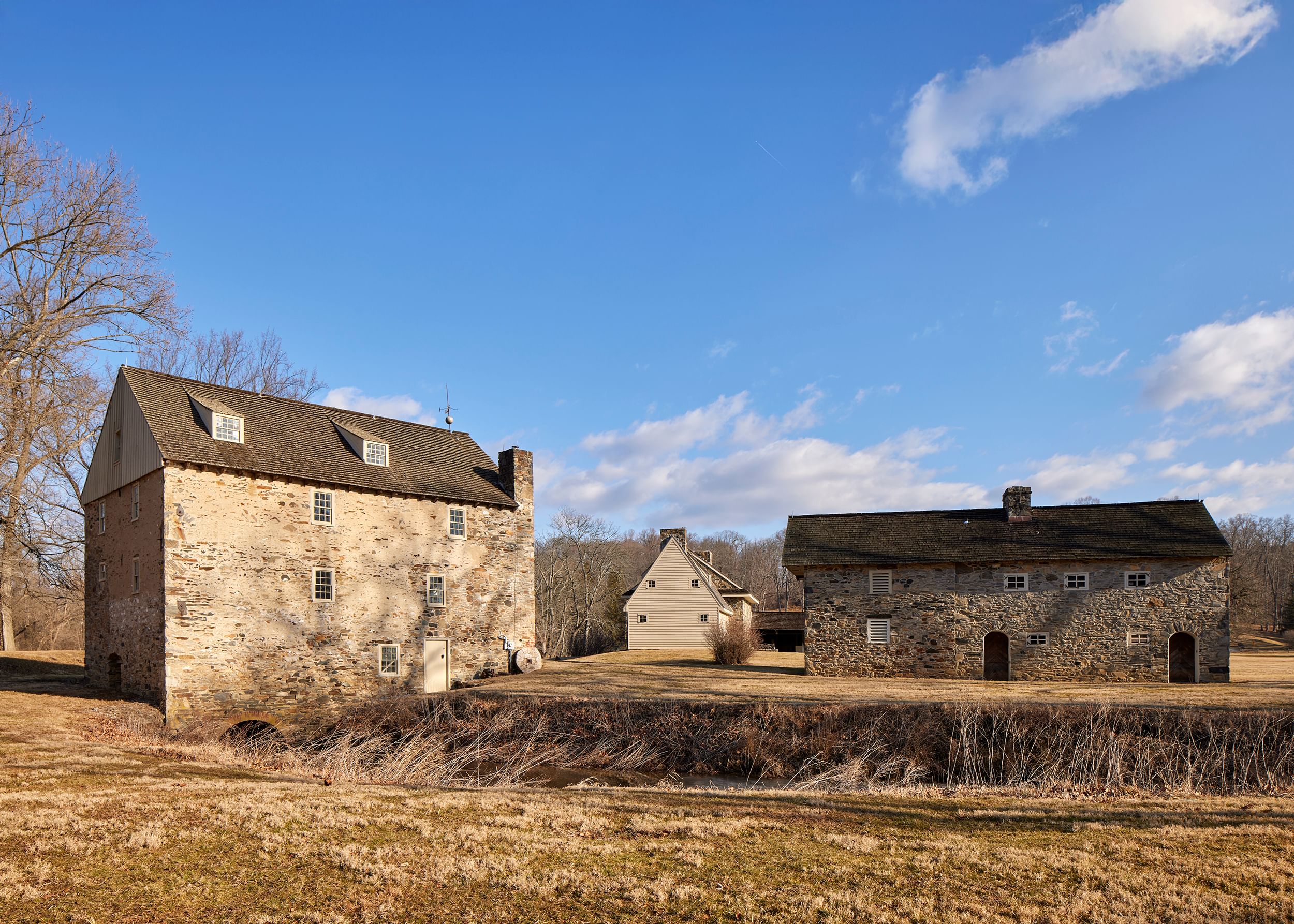 View of three large, rustic stone buildings on a bright, sunny winter day with brown grasses and bare trees.