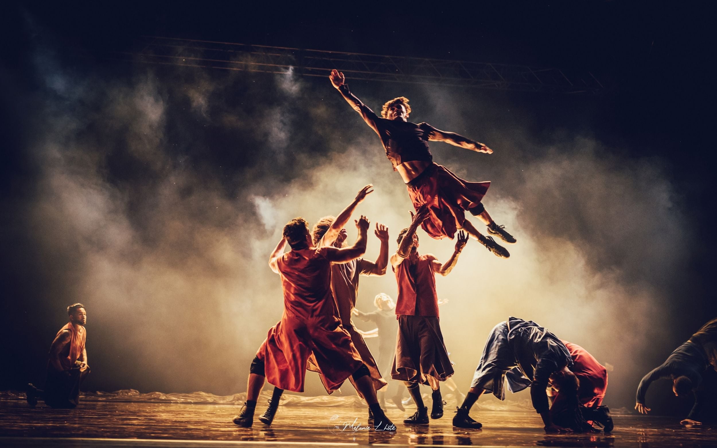 Six male dancers in red and blue outfits on a foggy stage, with one dancer in mid-air and three arms raised to catch him.