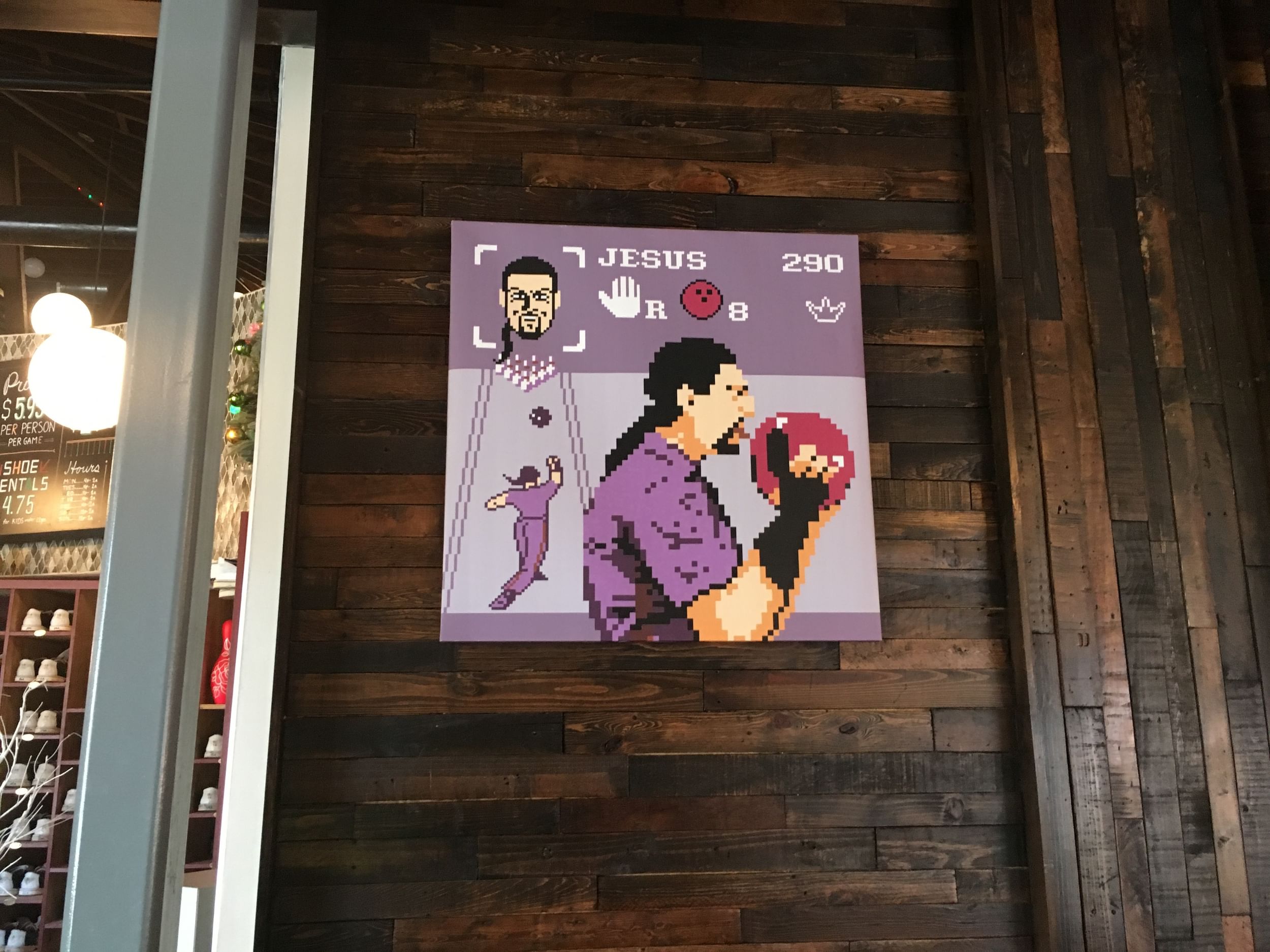 A photo of a pixelated image on a canvas on a wall of the Dude from The Big Lebowsky in a bowling alley