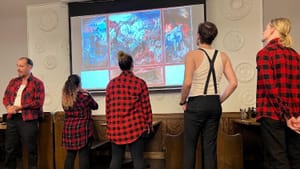 5 actors, 4 in red plaid shirts, stand around a grim war-scene engraving by Otto Dix, projected on the white wall of a bar