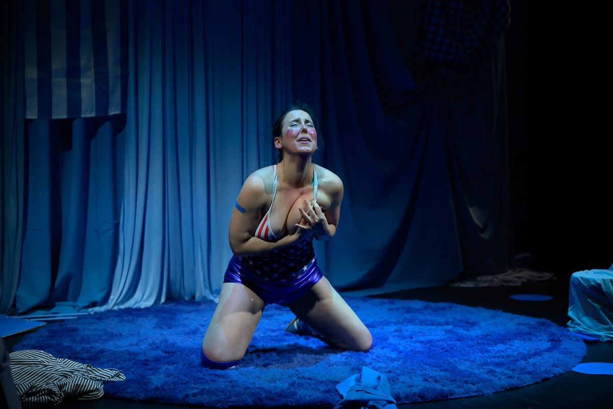 Minora, in a stars & stripes leotard, cries dramatically while kneeling on a blue rug, hands squishing her left breast.