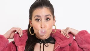 Minora, a white woman with brown hair wearing gold hoop earrings and pink plush jacket, blows a bubble with her gum.