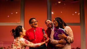 A scene from the play. 4 actors joyfully share a microphone, karaoke-style. One holds a baby. They’re behind a yellow couch