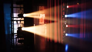 Backstage at a large theater: large white, red, and blue stage lights shine from the side through misty air.