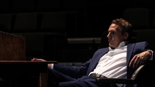 Actor Ben Jalosa Williams, a white man, plays William F. Buckley Jr. He wears a dark suit and lounges at a wooden desk.