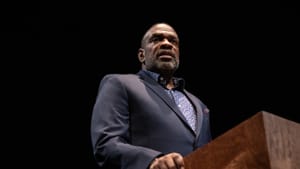 Actor Greig Sargeant, a Black man, plays James Baldwin. He wears a dark blazer and stands at a wooden lectern.